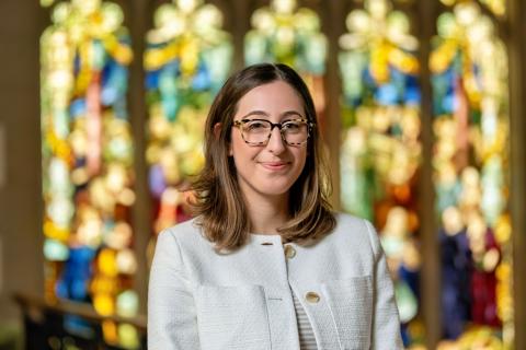 Stephanie Jackvony with shoulder-length brown hair and tortoiseshell glasses smiles while standing indoors, wearing a white textured jacket, with colorful stained-glass windows blurred in the background.