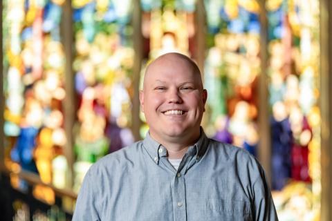Portrait of a person with a bright smile, standing in front of a colorful stained glass window.