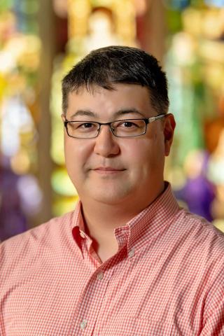 A person with short, dark hair and glasses smiles faintly at the camera. The person is wearing a red and white gingham collared shirt. The background is an abstract blur of warm, bright colors, suggesting a soft focus on stained glass or a vibrant natural setting.