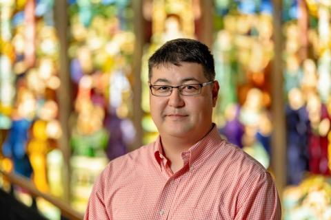 A person with short, dark hair and glasses smiles faintly at the camera. The person is wearing a red and white gingham collared shirt. The background is an abstract blur of warm, bright colors, suggesting a soft focus on stained glass or a vibrant natural setting.
