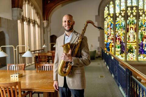 Joseph North in a tan suit stands in a library holding a saxophone. He is smiling, with wooden tables, brass lamps, and a large stained glass window visible in the background.