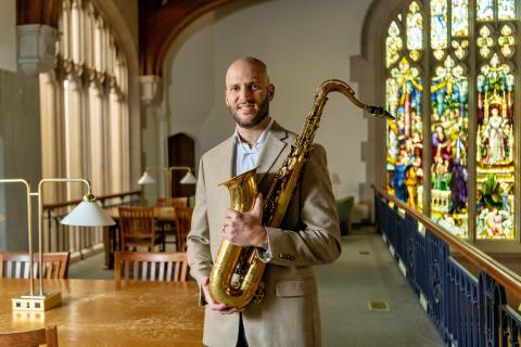 Joseph North in a tan suit stands in a library holding a saxophone. He is smiling, with wooden tables, brass lamps, and a large stained glass window visible in the background.