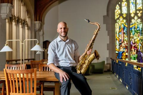 Joseph North in a white dress shirt sits on a wooden table holding a saxophone, smiling at the camera in a library with wooden chairs, brass lamps, and a large stained glass window in the background.