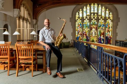 Joseph North in a white dress shirt sits on a wooden table holding a saxophone, smiling at the camera in a library with wooden chairs, brass lamps, and a large stained glass window in the background.