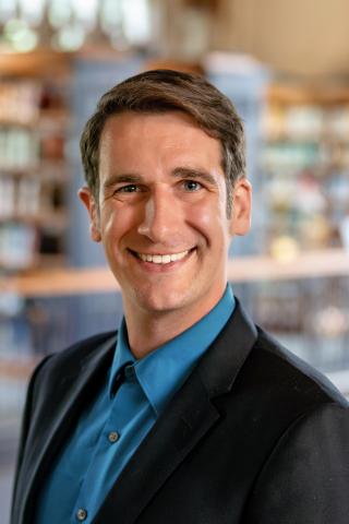 Maxwell Fuerderer wearing a blue shirt and dark blazer smiles while standing indoors, with blurred bookshelves in the background.