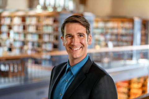 Maxwell Fuerderer wearing a blue shirt and dark blazer smiles while standing indoors, with blurred bookshelves in the background.