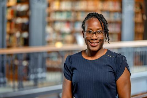 Kimberly Williams Brown wearing glasses and a navy blue pinstriped top smiles while standing indoors, with blurred bookshelves in the background.