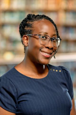 Kimberly Williams Brown, head turned to the side, wearing glasses and a navy blue pinstriped top smiles while standing indoors, with blurred bookshelves in the background.