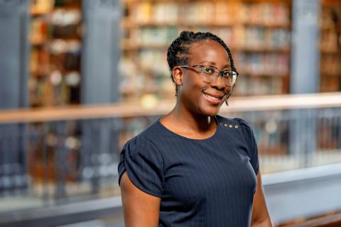 Kimberly Williams Brown, head turned to the side, wearing glasses and a navy blue pinstriped top smiles while standing indoors, with blurred bookshelves in the background.