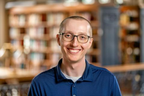 Jacob Erickson smiling with glasses in a blue polo shirt, posing in front of a blurred bookshelf background.