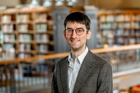 Tommasi Pier Carlo smiling with glasses, wearing a brown plaid blazer and white shirt, with a blurred library background.