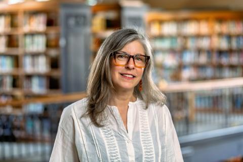 Erin McCloskey, with shoulder-length gray hair and glasses, smiling in front of bookshelves in a library.