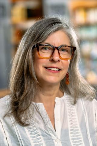 Erin McCloskey, with shoulder-length gray hair and glasses, smiling in front of bookshelves in a library.