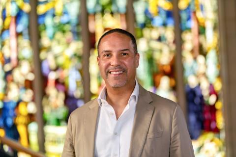 Salvador Contes, smiling with short dark hair and a neat goatee. He is wearing a tan suit jacket over an open-collared white shirt, set against a vibrant, softly blurred background of colorful stained glass.