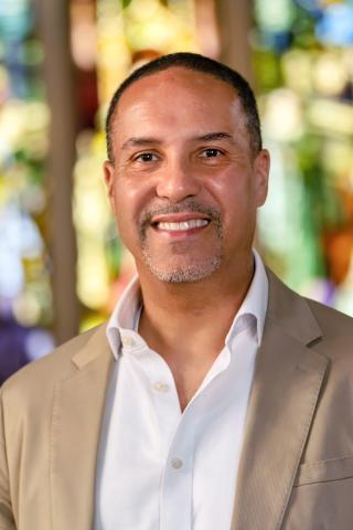 Salvador Contes, smiling with short dark hair and a neat goatee. He is wearing a tan suit jacket over an open-collared white shirt, set against a vibrant, softly blurred background of colorful stained glass.