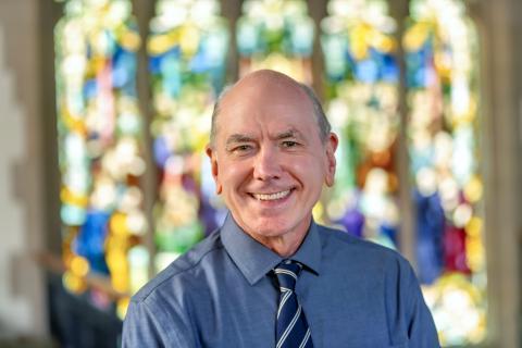 Peter Lemieszewski, wearing a blue shirt and striped tie, with stained-glass windows in the background.