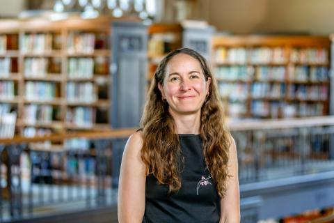 Colette Salyk, with long, wavy brown hair and gold hoop earrings smiling, wearing a black top with subtle pink floral embroidery in front of a soft-focused library background filled with wooden bookshelves.