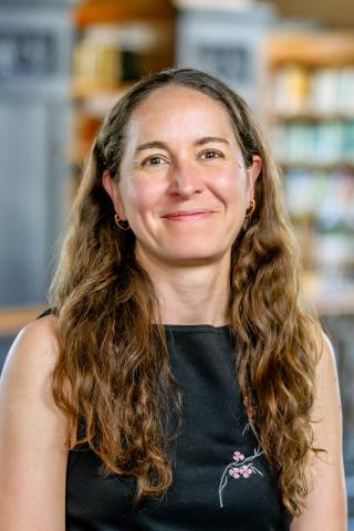 Colette Salyk, with long, wavy brown hair and gold hoop earrings smiling, wearing a black top with subtle pink floral embroidery in front of a soft-focused library background filled with wooden bookshelves.