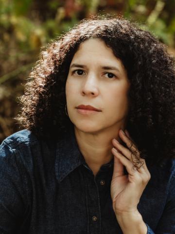 A person with dark, shoulder-length curly hair wears a dark blue denim button-up shirt and rests one hand against their neck while looking forward. The background is a softly blurred outdoor setting with natural sunlight.
