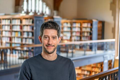 Jesse Delia smiling with short dark hair and light stubble, wearing a plain dark crewneck shirt in a library with wooden bookshelves blurred in the background.