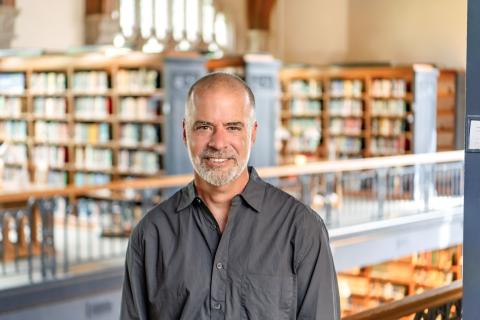 Chris Grove with short hair and a graying beard, wearing a dark gray button-up shirt set against a softly blurred library background with wooden bookshelves.