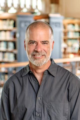 Chris Grove with short hair and a graying beard, wearing a dark gray button-up shirt set against a softly blurred library background with wooden bookshelves.