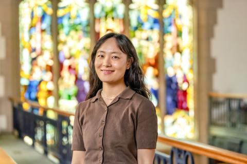 Soo Jin Jeon with long dark wavy hair, wearing a brown short-sleeved button-up knit shirt and a delicate necklace, set against a softly blurred background of a colorful stained-glass window.