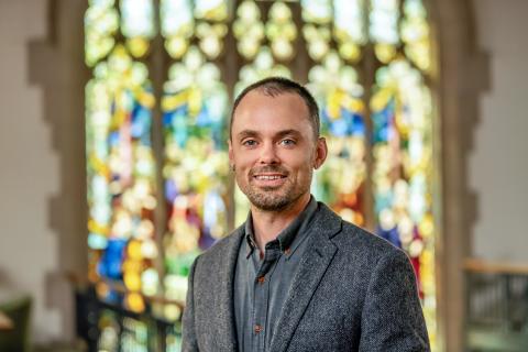 John Garrard Stehlin smiling with short dark hair, light facial hair, and blue eyes, wearing a textured gray blazer over a dark button-down shirt against a softly blurred stained-glass window background.