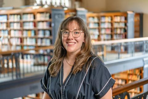 Maya Marie Van Nuys smiling, long wavy brown hair with bangs, and clear-framed glasses, wearing a black top with white piping in front of a softly blurred library background with wooden bookshelves.