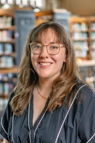 Maya Marie Van Nuys smiling, long wavy brown hair with bangs, and clear-framed glasses, wearing a black top with white piping in front of a softly blurred library background with wooden bookshelves.