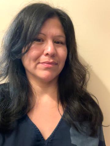 Leora Gansworth with long dark wavy hair, wearing a dark V-neck top and smiling gently against a light neutral wall.