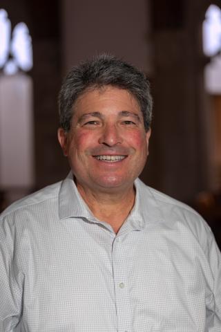 Marc Smith smiling, wearing a light gray open-collar shirt with a micro-grid pattern, against a softly blurred interior background.