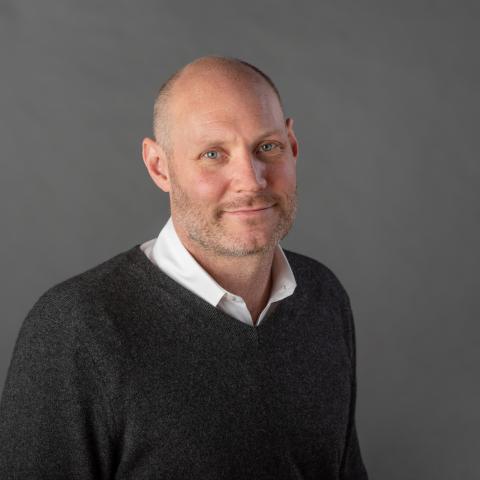 Hadley Creighton Bergstrom wearing a white collared shirt and dark gray sweater against a gray background.