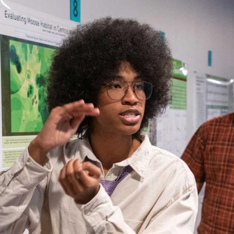 Student standing in front of a poster explaining it while gesticulating to a group of people off camera