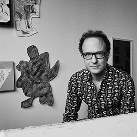 poet Wayne Koestenbaum seated in front of a table with abstract art works on the wall behind him.