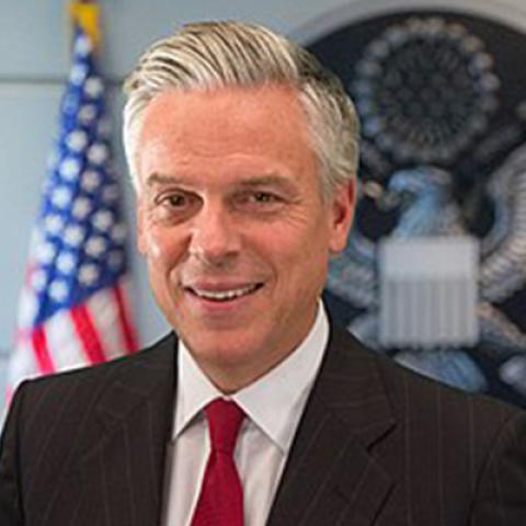 an official portrait of former U.S. Ambassador Jon M. Huntsman Jr., who is wearing a jacket and tie and standing in front of an American flag behind him.