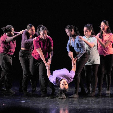 Seven women on a stage holding one of them in a dance position