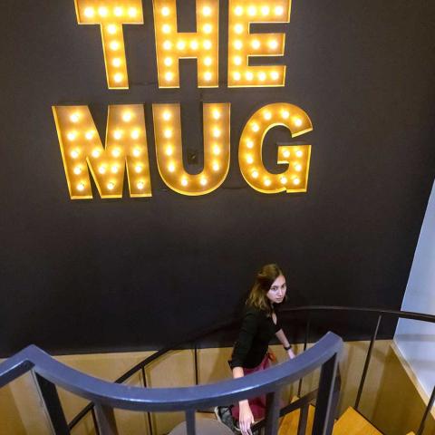 A person with long brown hair walks up a spiral staircase, underneath giant letters with lightbulbs that spell "The Mug."