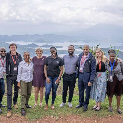 Nine people stand in a row on a grassy hilltop, smiling at the camera. Behind them is a view out over mountains and a river. The sky is filled with rolling clouds.