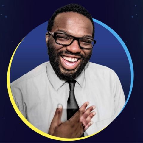 Picture of composer Shavon Lloyd; smiling wearing gray collared shirt with a black tie, hold his hand over his heart.