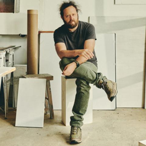 Noah Kalina seated on a stool in an art studio.