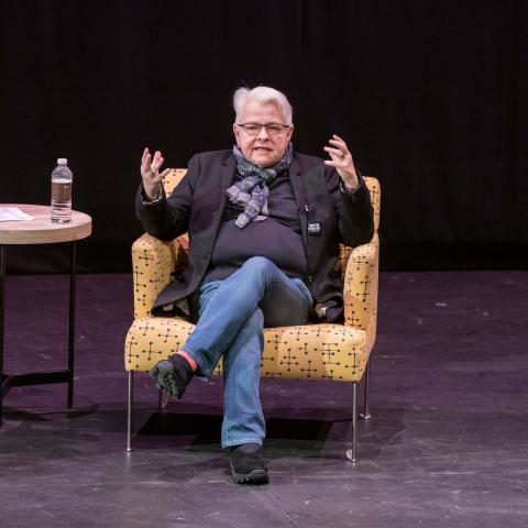 Pulitzer Prize-winning playwright Paula Vogel wearing a black shirt and jacket and jeans sitting in a chair in the Martel Theater