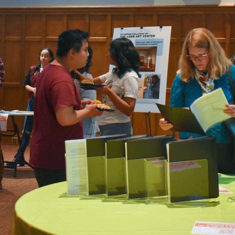 Standing person reading a printed presentation in a folder and others in the background looking at poster presentations on easels