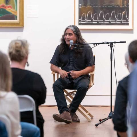 Person seated behind a microphone in an art gallery facing the camera and speaking to a seated audience