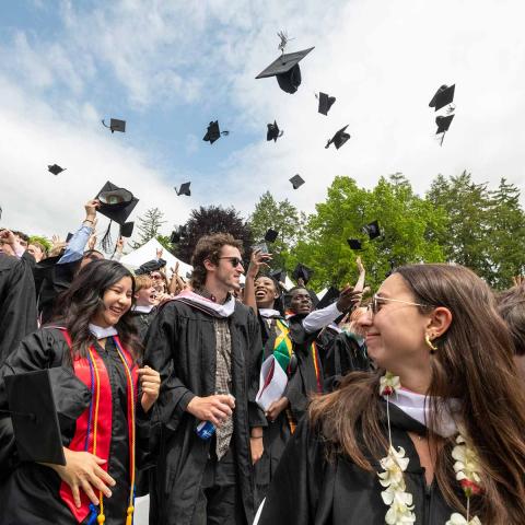 A group of people wearing dark graduation robes stand outside on a sunny day, tossing their hats into the air.