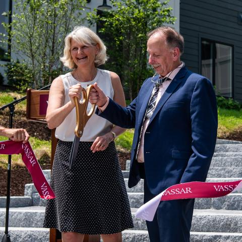 Man and woman holding giant scissors cutting a red ribbon with "Vassar" printed on the ribbon.