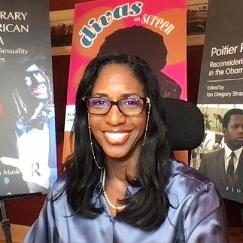 Mia Mask wearing a satin blouse, pearl necklace, and big smile posed in front of three large posters featuring covers of books she has worked on: Contemporary Black American Cinema, Divas on Screen, and Poitier Revisited.