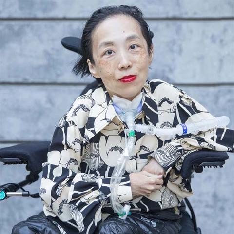 Portrait of Alice Wong sitting in a wheelchair with a breathing tube attached to her throat.