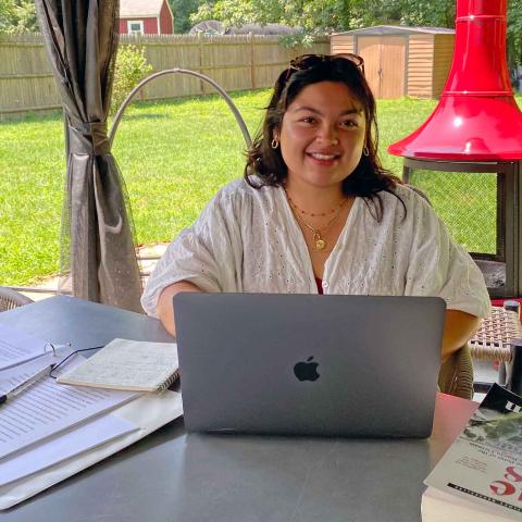 Person sitting at a table on a porch in the shade with a laptop, writing pad and a book smiling