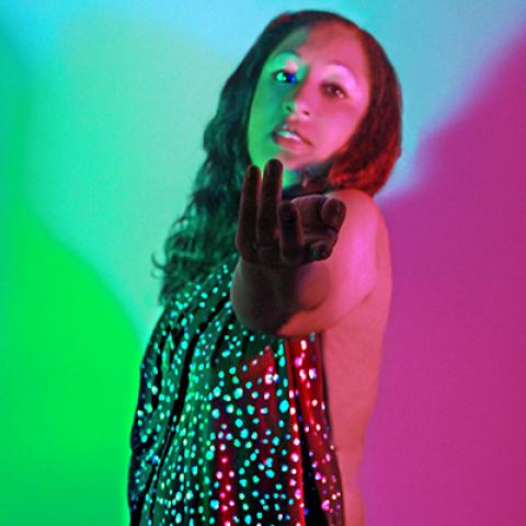 A women in a sparkly gown stares at the camera while holding her hand out in a beckoning pose.